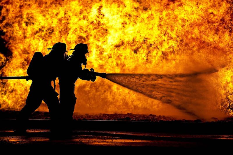Silhouette de deux pompiers devant un mur de feu arrosant les flammes.